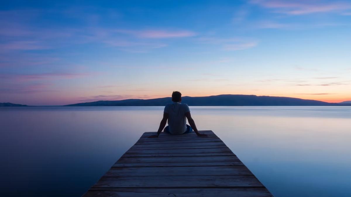 A man sits alone at the end of a wooden dock at dusk, facing a still lake