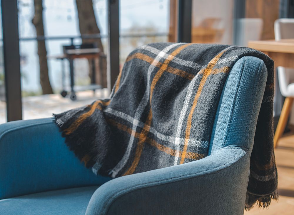 A soft blue armchair with a grey and mustard plaid wool throw over it, beside a window